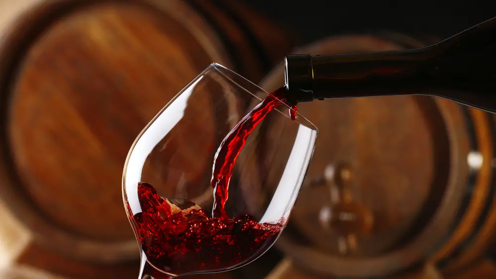 Pouring red wine from bottle into glass with wooden wine casks on background