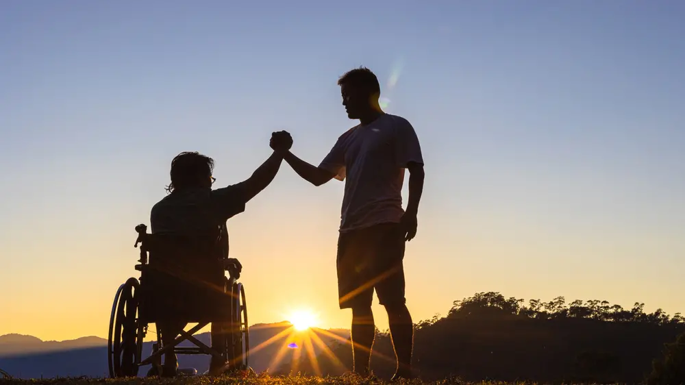 Silhouette of joyful disabled man in wheelchair raised hands with friend at sunset , Shutterstock