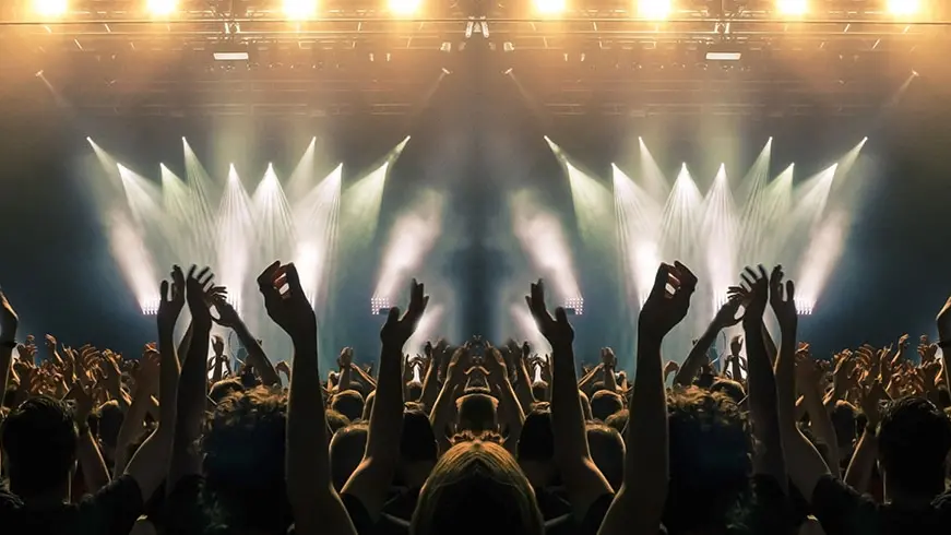 A shot taken in front of a concert stage lit in the night, people are visible waving and clapping, but no one is recognizable. , Getty Images/iStockphoto