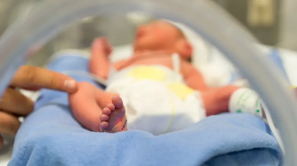Photo of a premature baby in incubator. Focus is on his feet and toes. The doctor is touching him to check his reflexes. There are cables and tubes in the out-of-focus area. , Getty Images